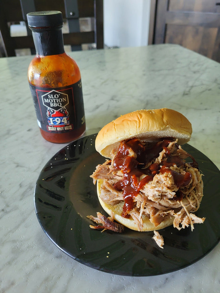 Pulled pork sandwich on a bun with barbecue sauce, next to a bottle of Slo-Motion BBQ I94 barbecue sauce.